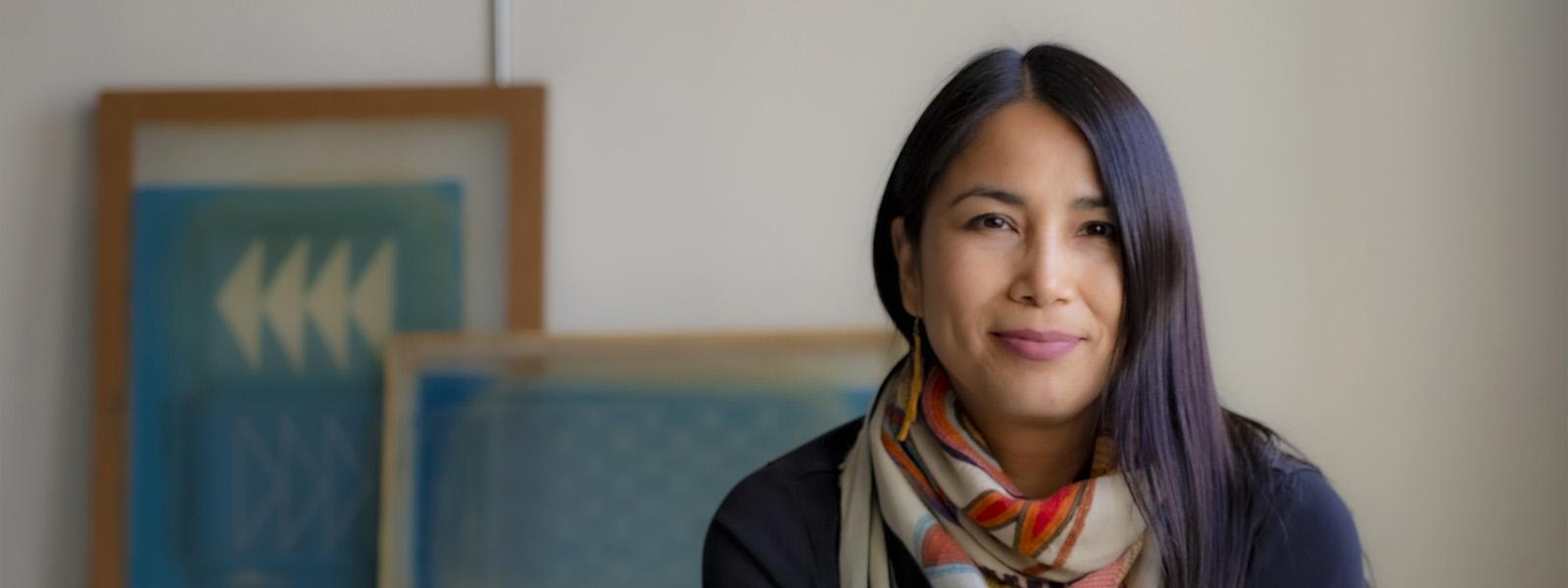 A headshot of a woman with long, black hair. She wears a grey scarf with a red, floral pattern on it and a dark long sleeved top. Behind her is a white wall with two framed art pieces that consist of light blue and white patterns.