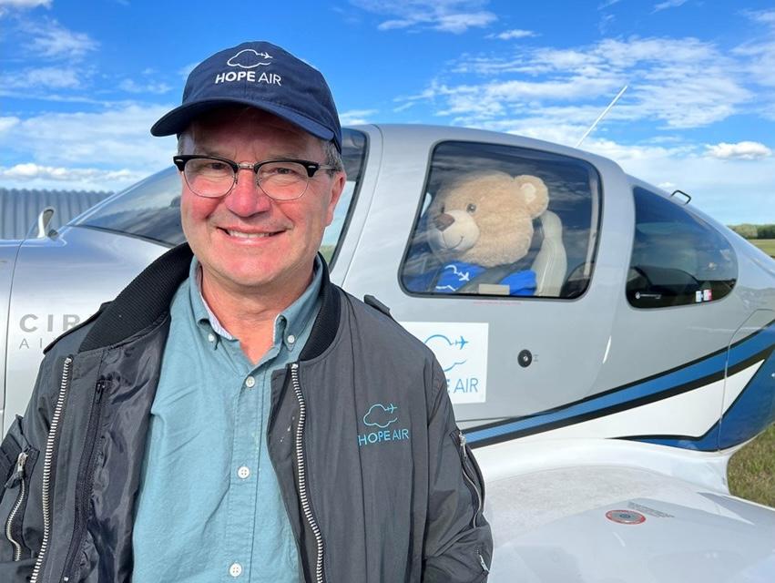 A man wearing a light blue shirt with a grey jacket over it stands in front of a small jet plane. In the pilot's seat of the plane is a big stuffed teddy bear.