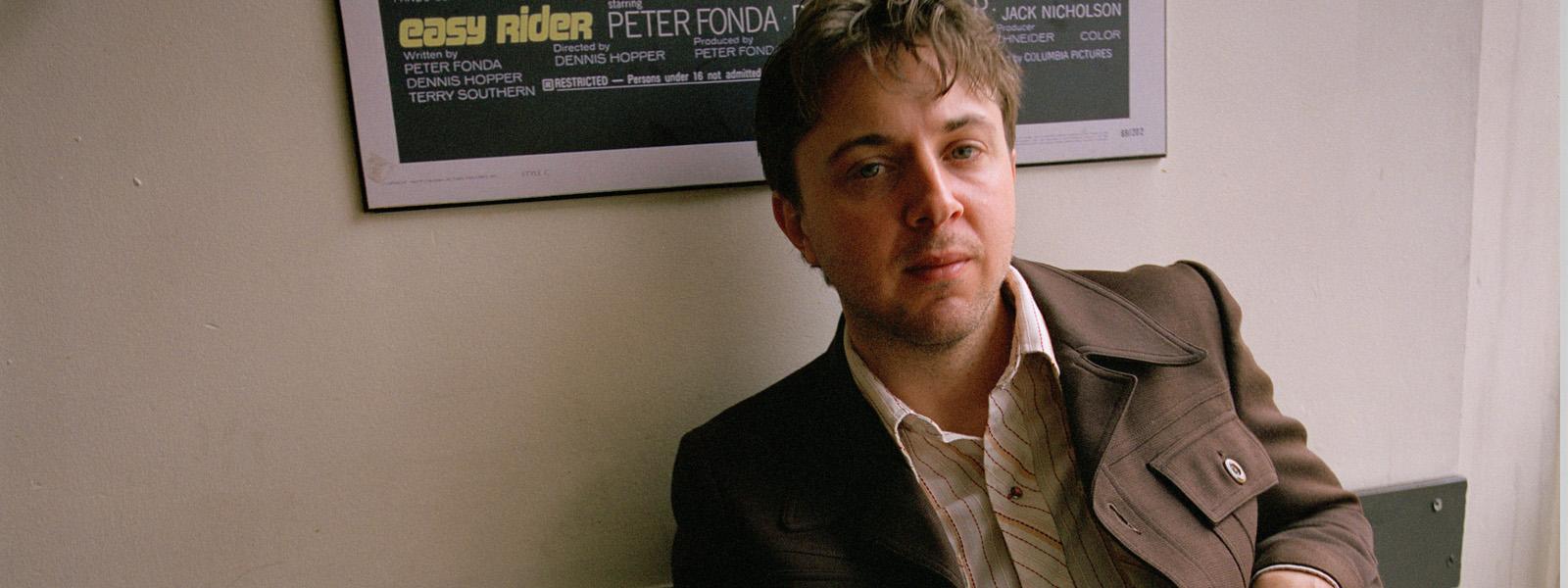 A man with short brown hair sits in front of a beige wall wearing a brown blazer and a white plaid collared shirt underneath.