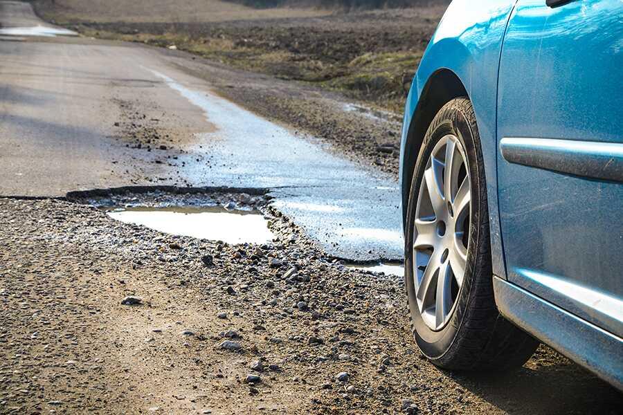 A blue car on a dirt road with a pothole in the road.