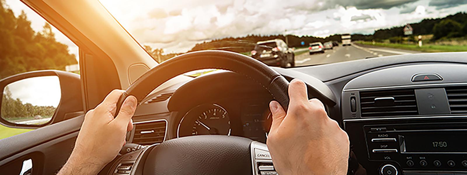 A driver’s point of view. Two hands hold a black steering wheel securely. Out of the windshield, a highway is visible with trees lining both sides. There is minimal traffic on the road, and the sun is shining.
