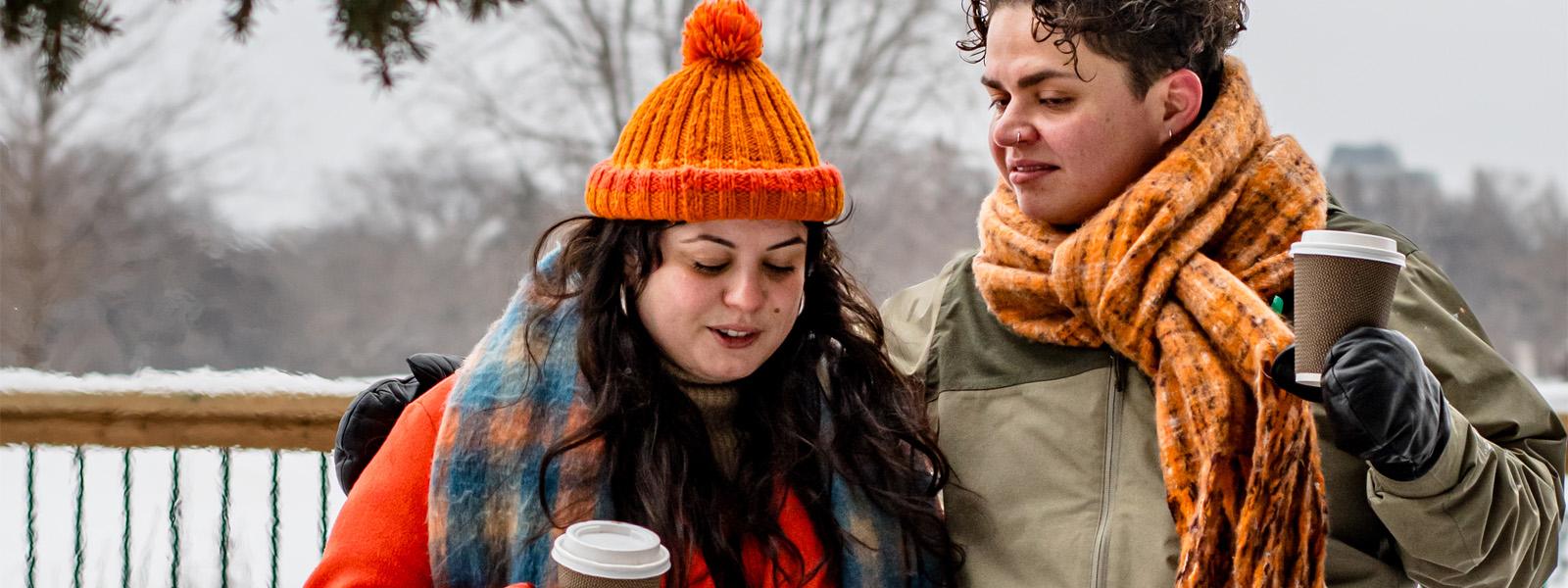 Two people wearing jackets, mitts and scarves stand outdoors with coffee cups in their hands. Behind them is a field of snow and a forest.