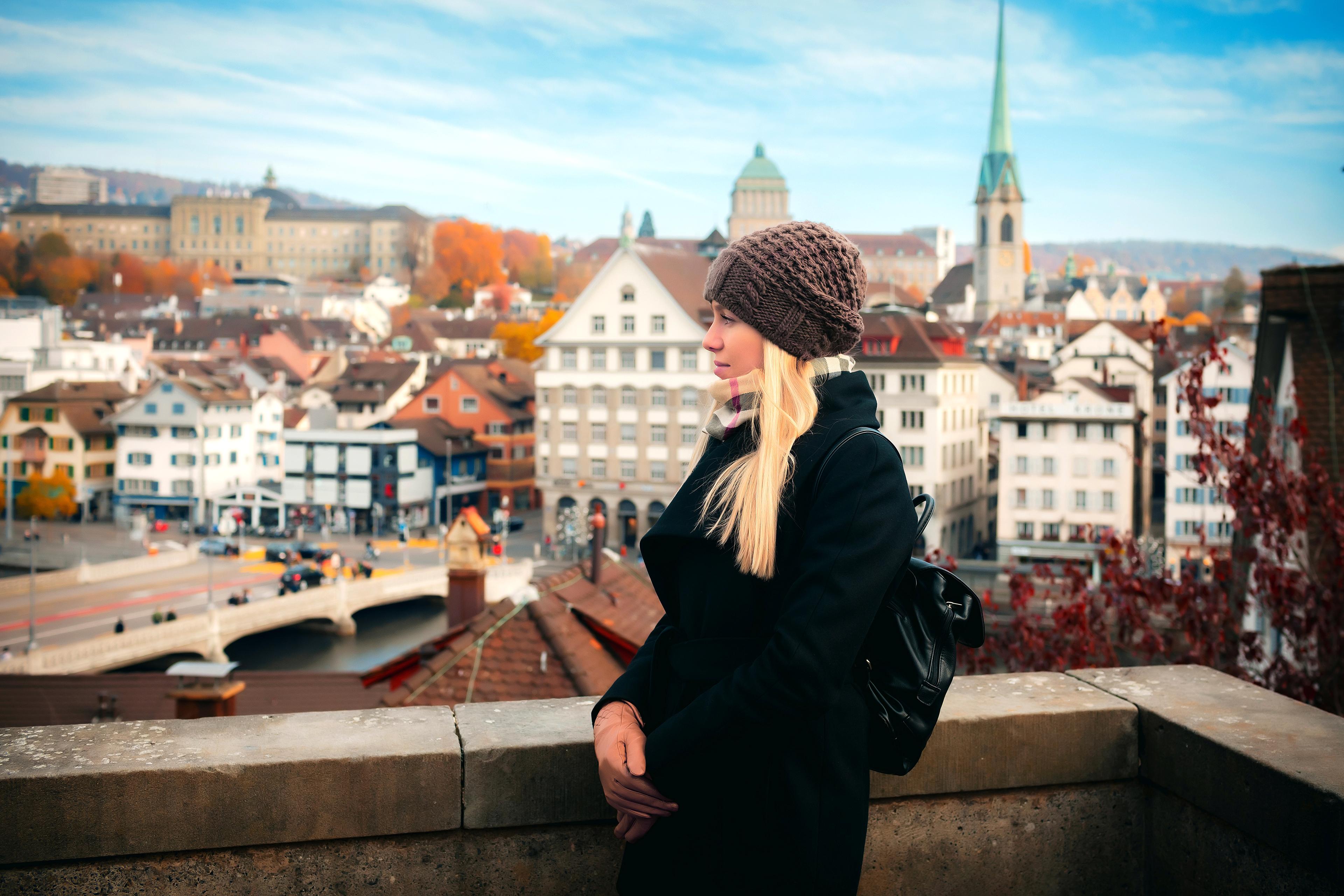 A woman wearing a black jacket and a brown beanie stands beside the edge of a rooftop. Behind her is a city, with many trees sporting fall-coloured leaves.