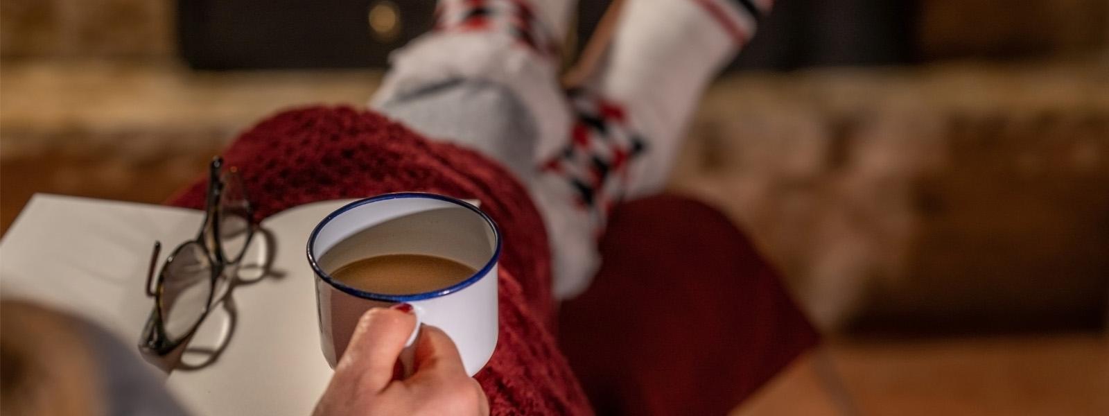 The lap of a person sitting indoors by a fireplace. They have a mug of coffee in their hands, with a book open and a pair of black glasses on top. Covering their legs is a red, knitted blanket.