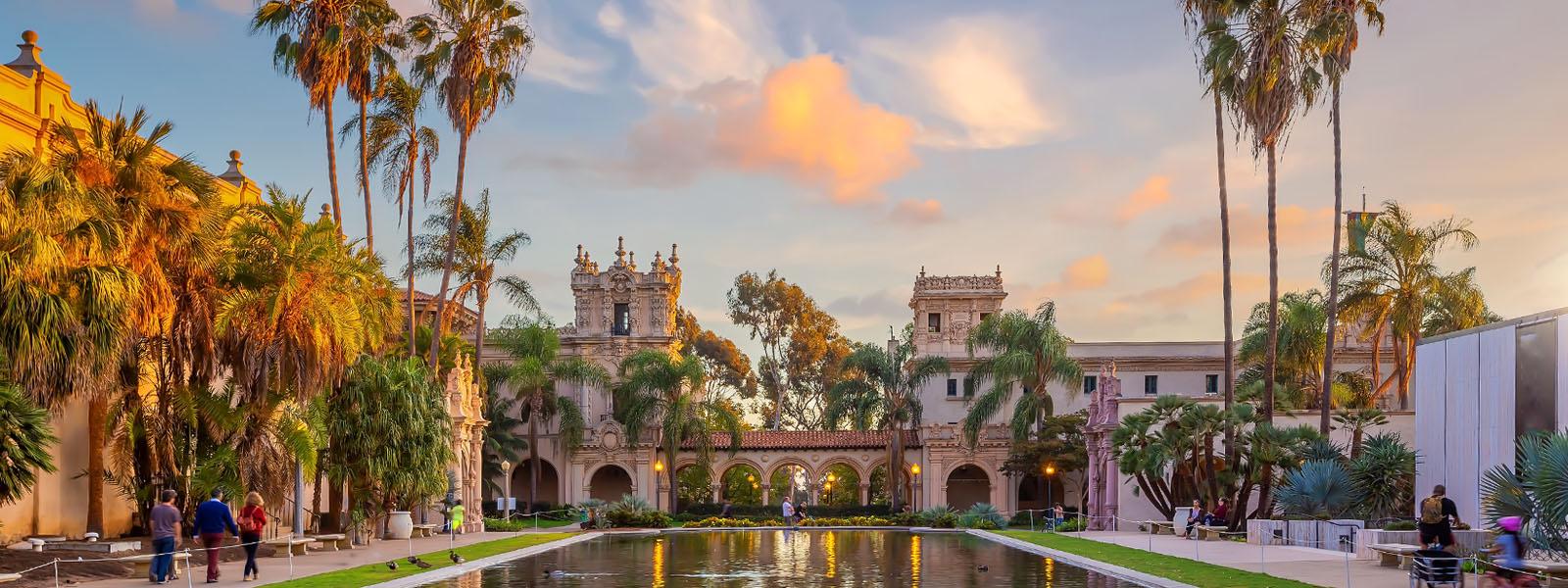 An outdoor plaza with a large pool in the centre. On the edges of the plaza are tall palm trees, and an a castle-like entrances with arches that line the bottom.