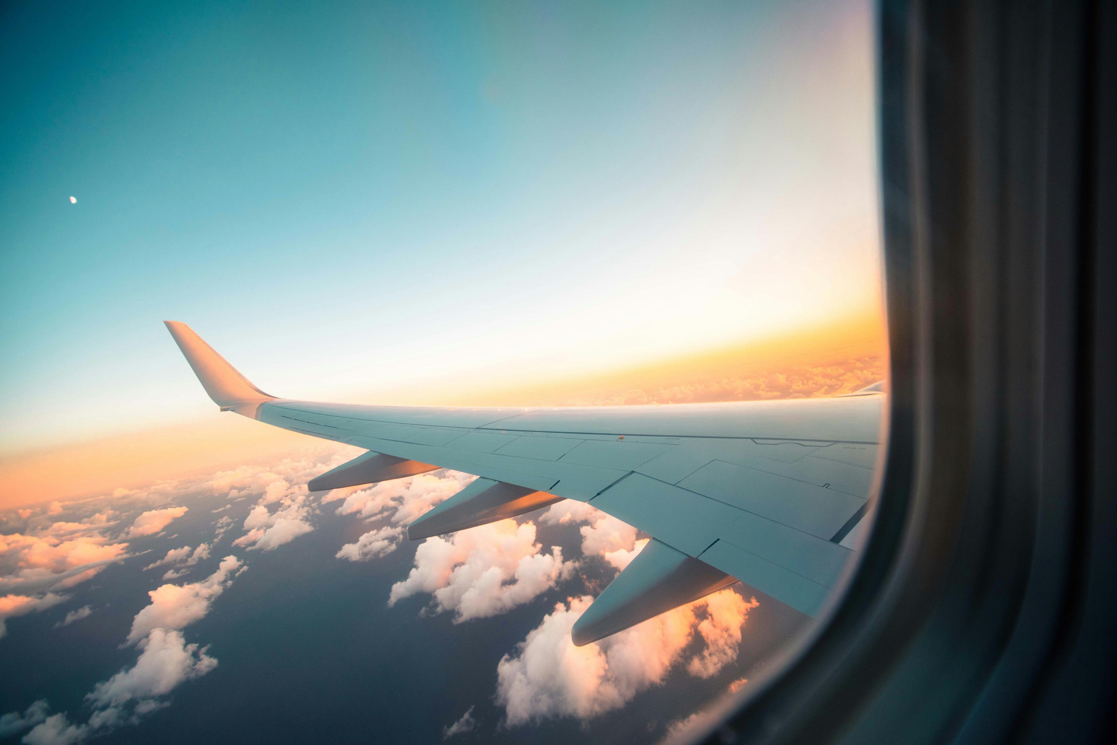 The view out of a plane window: the wing of the plan stretches outward into a blue sky with a setting sun, below the aircraft are clouds and ocean.