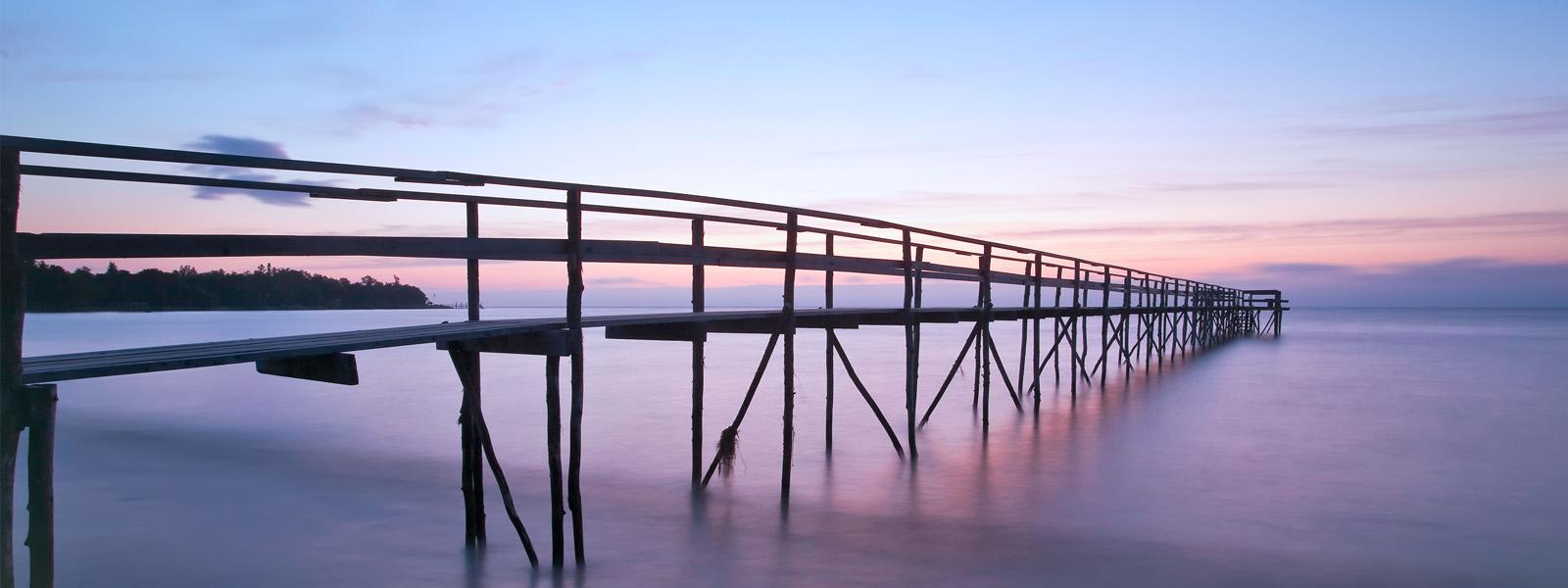 A minimalistic pier extends out from a white sand beach to the water. The sun is setting, and sky behind it is pink and purple.
