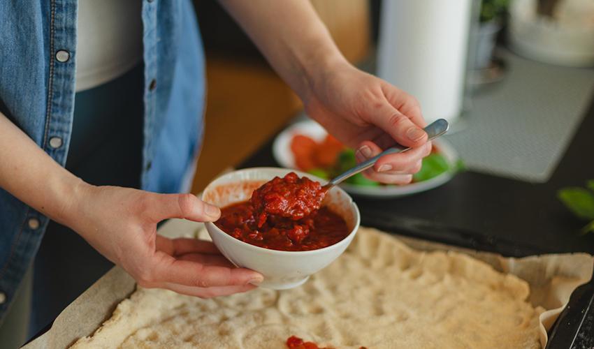 Someone spooning tomato sauce out of a bowl and spreading it on pizza dough.