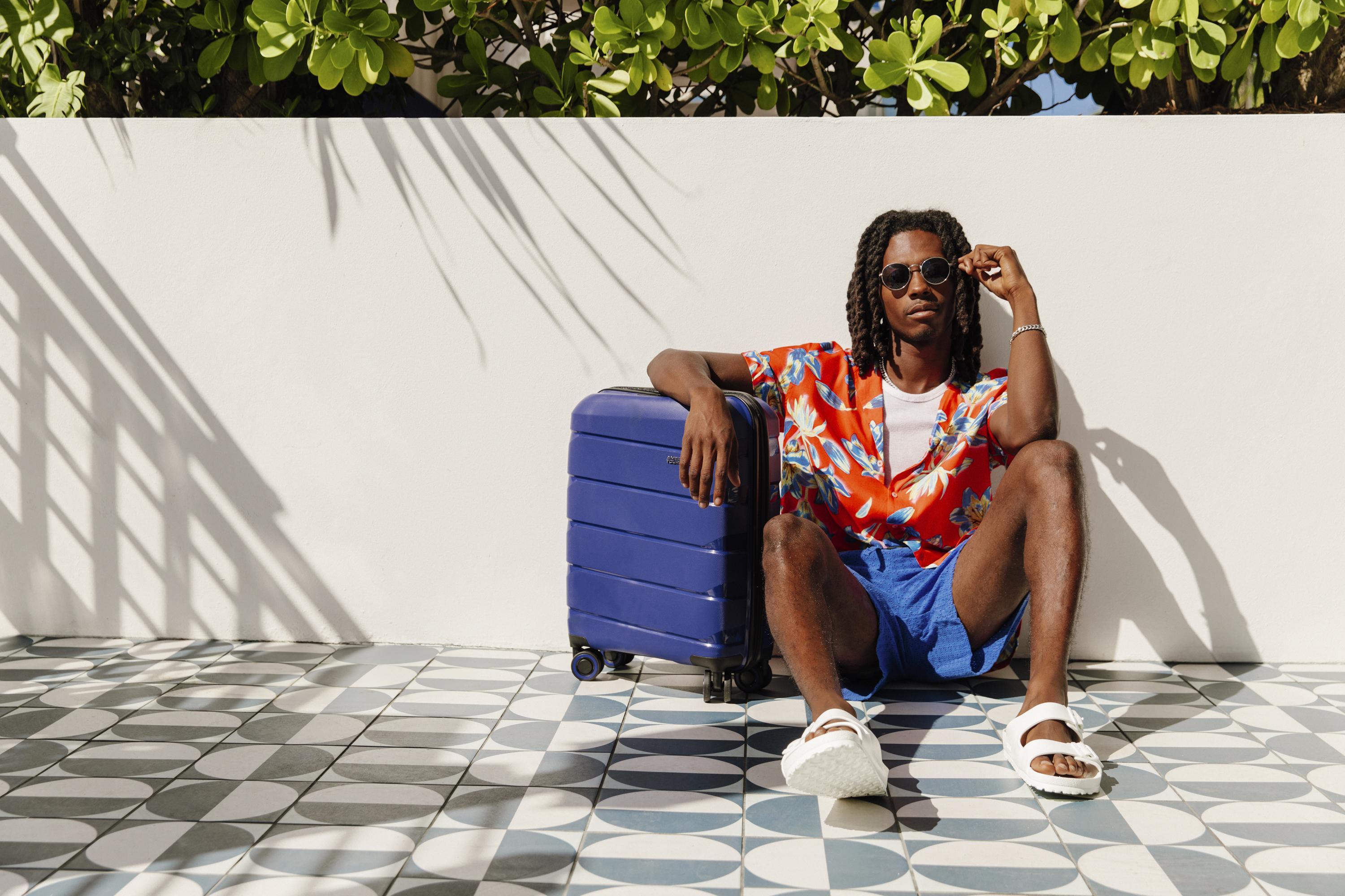 A man wearing a bright, floral button-up shirt and blue shorts sits on the ground on an outdoor patio next to a blue suitcase. He rests his arm on the suitcase with the sun shining down on him, and behind him are green leaves.