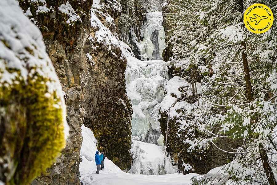 A hiker observing a frozen waterfall in a snow-covered forest canyon.