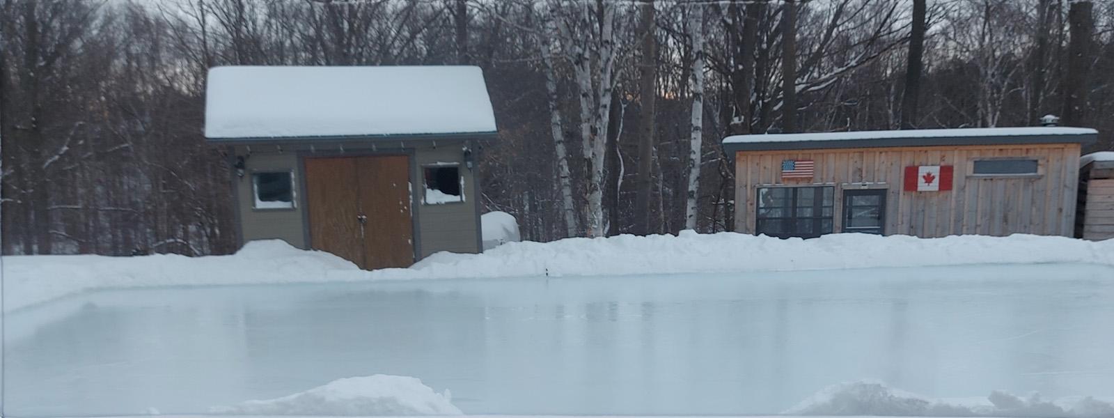 A backyard ice rink, surrounded by snow. Behind the ice are two sheds, one with a Canadian flag on the side, and a dense forest of dead trees.