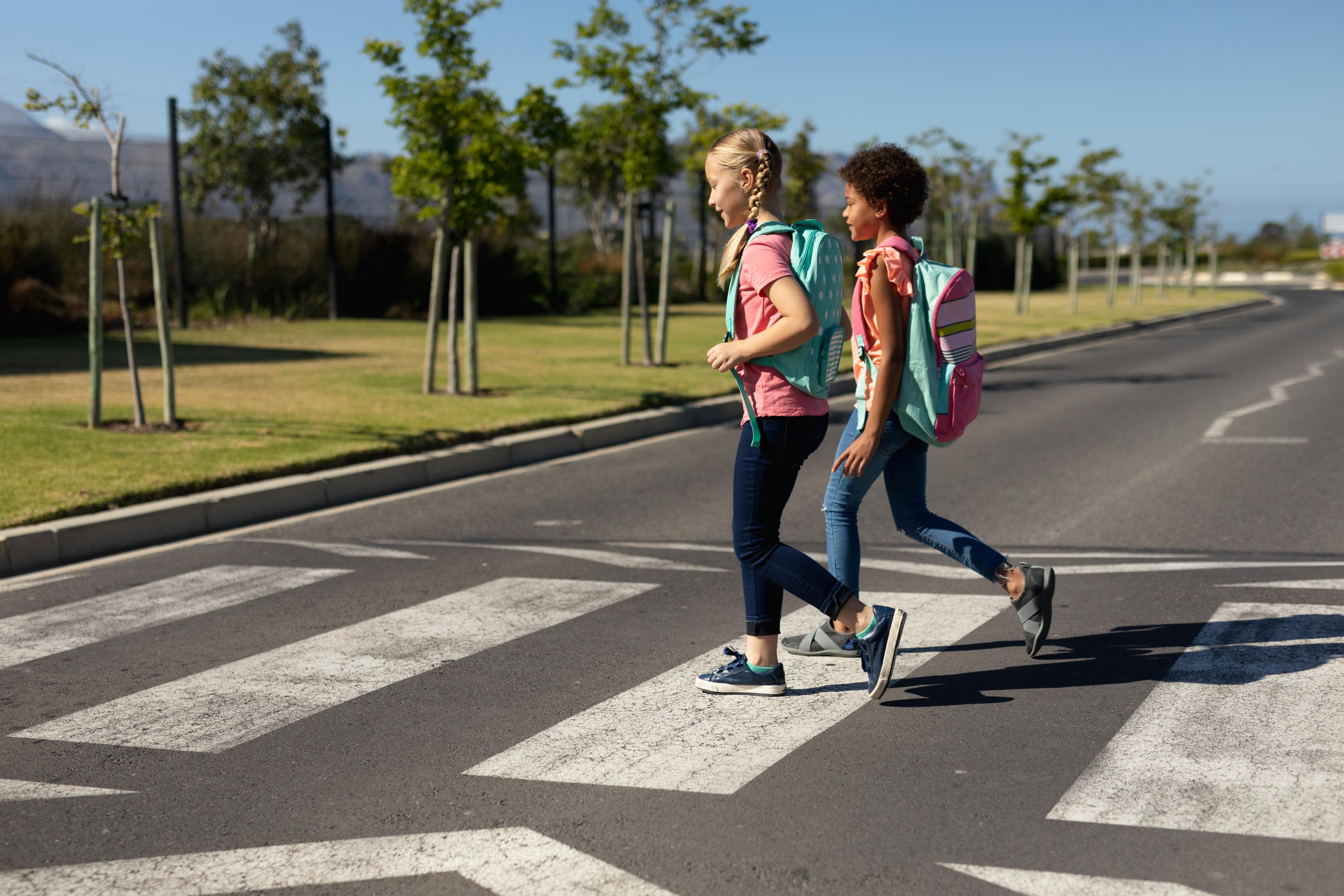 Two children wearing blue jeans and t-shirts, as well as brightly coloured backpacks, cross the road at a designated crosswalk.