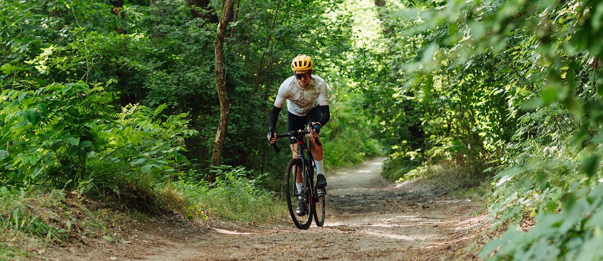 A biker, wearing a yellow helmet and a white t-shirt, rides his bike down a gravel trail through a dense forest, towards the camera.