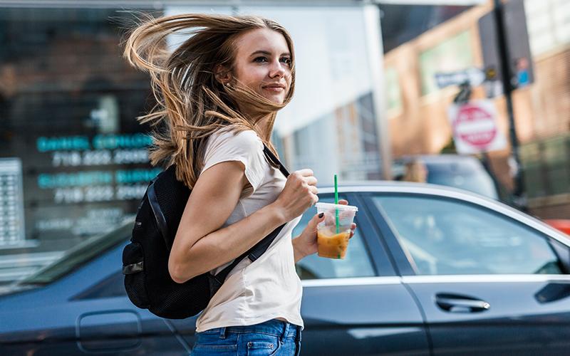 Woman holding an iced coffee walking on a sidewalk by a car