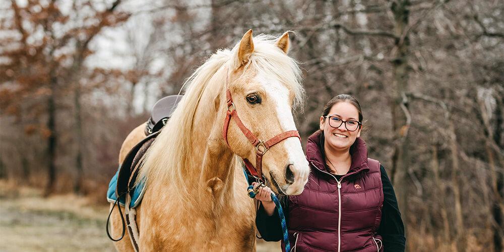 A woman smiling next to a palomino horse with a red halter, both standing outdoors.