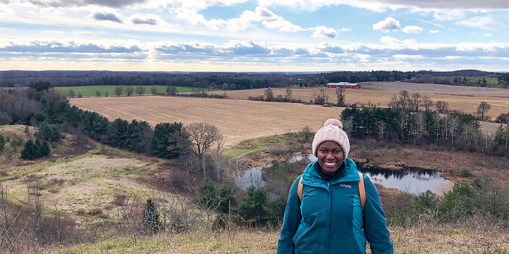 A person stands smiling in the foreground, wearing a winter hat and coat, with a scenic countryside landscape in the background.