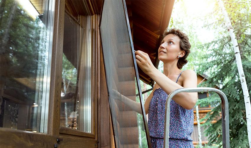 A woman smiles as she removes a window screen from outside a cottage to inspect it.