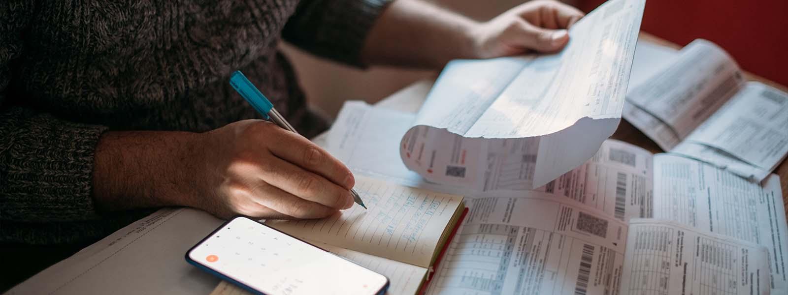 A close-up shot of a person sitting at a desk covered in paperwork. They are wearing a grey sweater with the sleeves slightly pulled up to keep their hands free as they hold a piece of paper in one hand while writing in a notebook with the other.