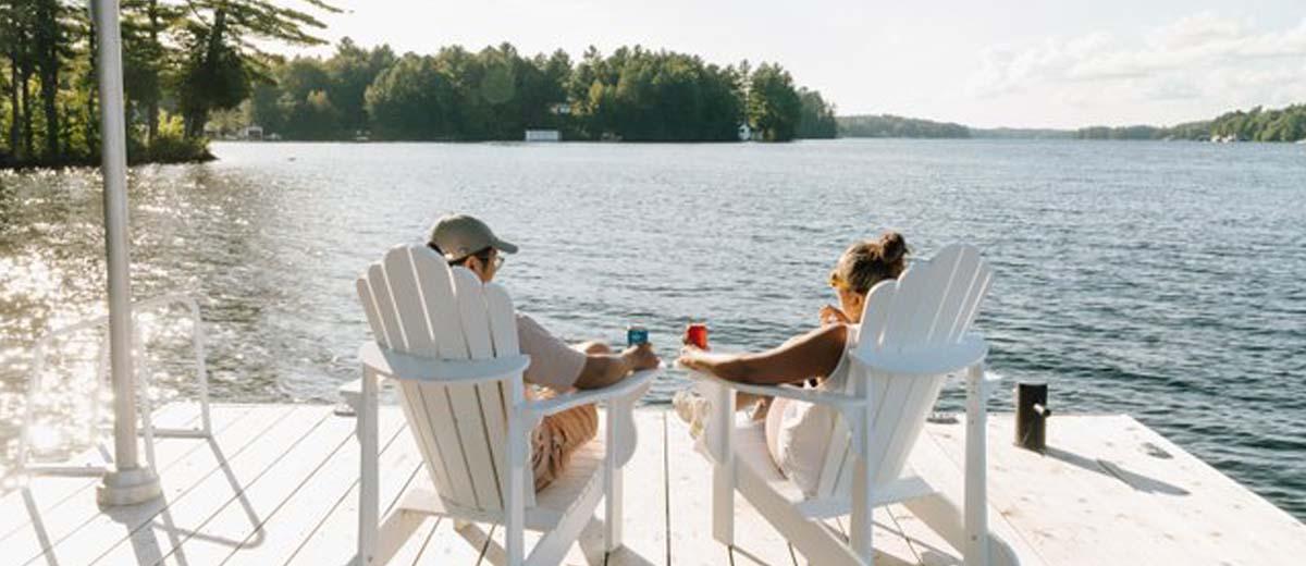 A view from behind of a man and woman sitting in white Muskoka chairs on a dock looking out at a lake.
