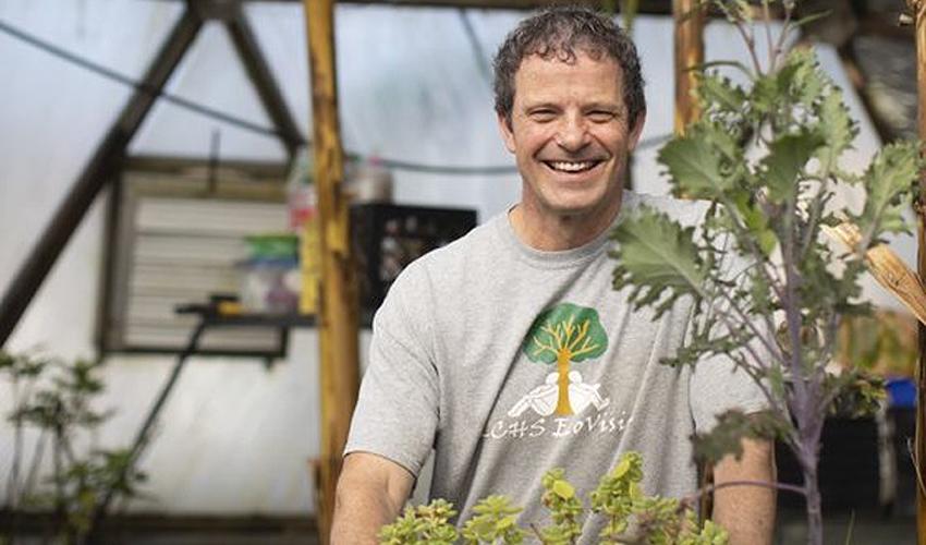 A man smiling in front of a greenhouse full of plants.