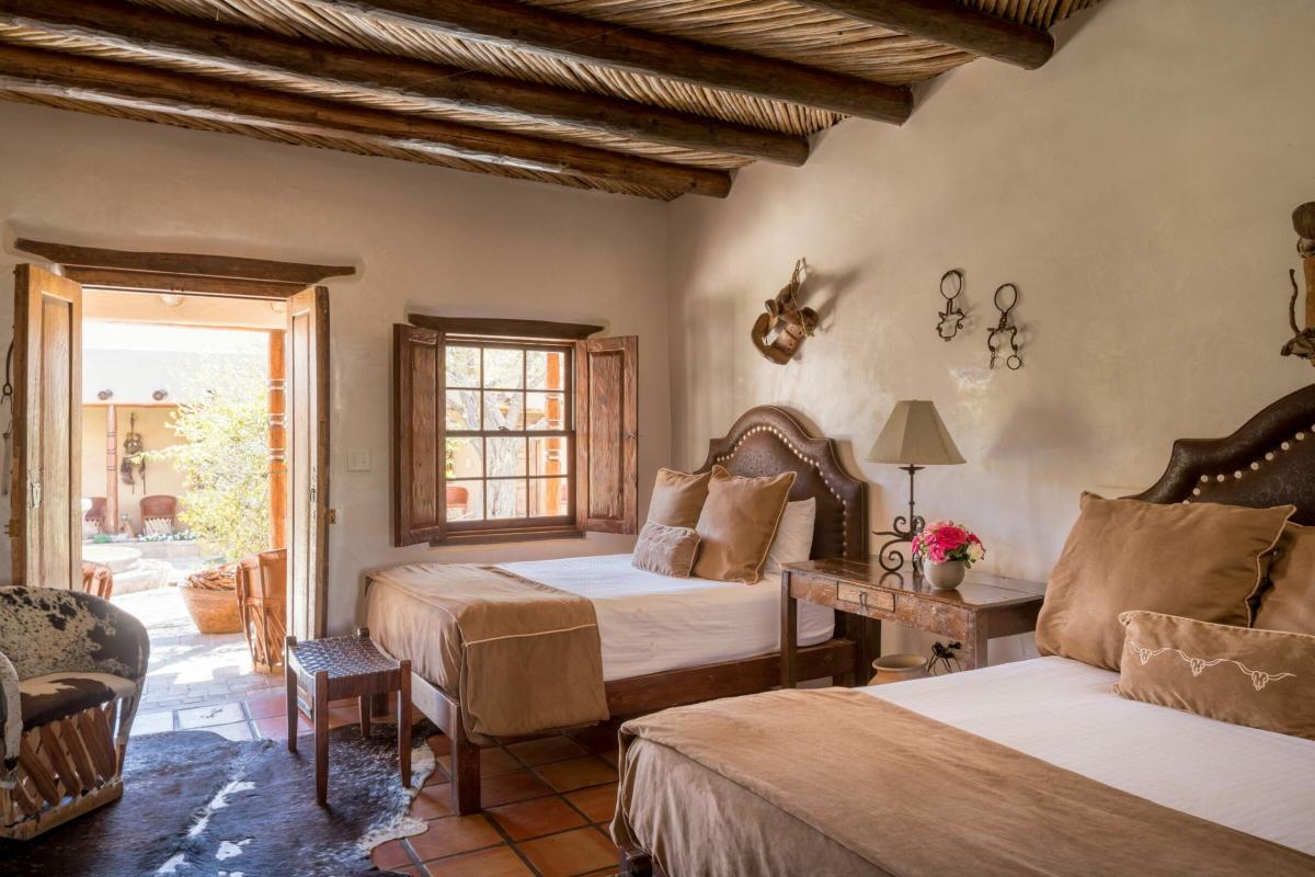 A hotel room with two beds and an open door leading to a garden. On the beds are brown pillows and white sheets, and between the two beds is a small table with a lamp.