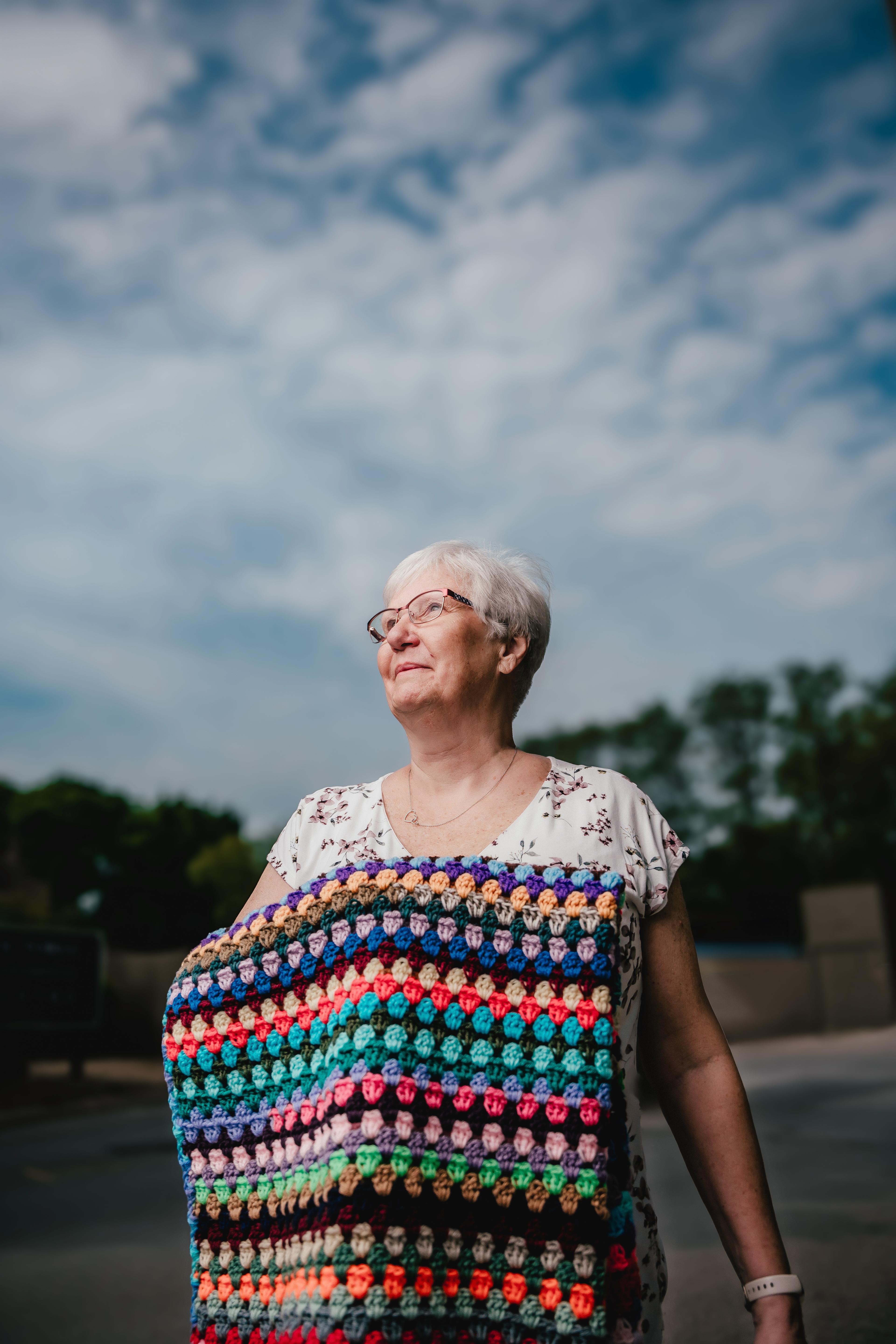 A woman with grey hair and a white t-shirt stands outside under a cloudy sky holding a rainbow-coloured crochet blanket.