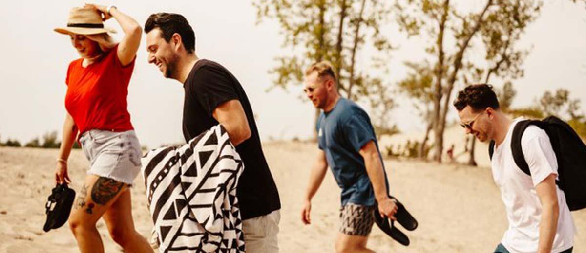 A woman in a straw hat and red T-shirt is walking with three men on a sandy beach. She is holding her shoes and so is one of the men. One is holding a beach blanket and another is wearing a backpack.