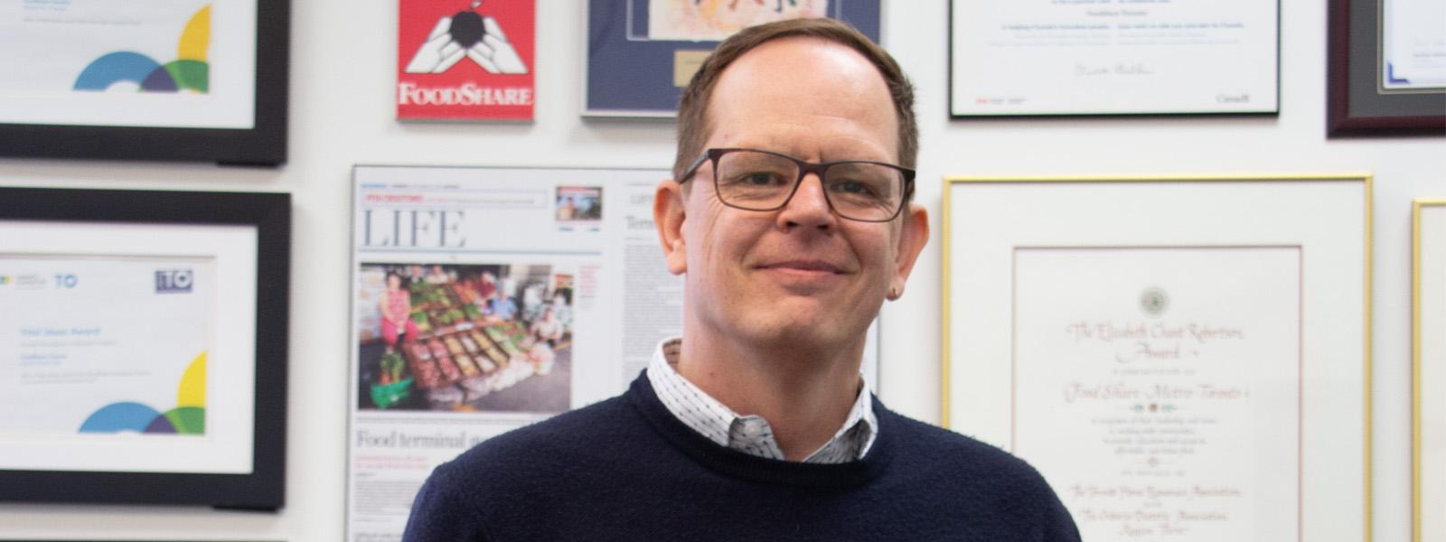 A man wearing black glasses and a collared white shirt under a black sweater, stands in front of a wall decorated with framed documents including a degree, pages from a magazine and artwork.
