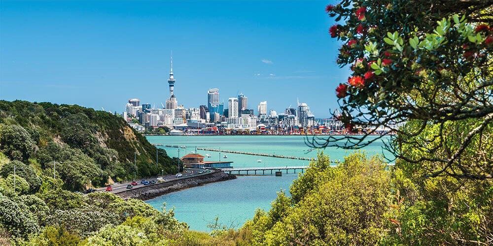 A scenic view of a city skyline with a pier extending into the ocean, framed by lush greenery on a sunny day.