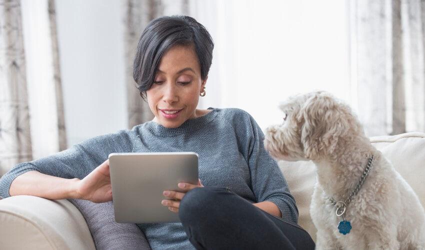 A woman sitting on a couch with her dog looking at a tablet.