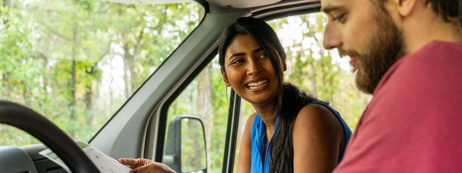 A white bearded and moustached man in a red T-shirt is sitting in the driver’s seat of a parked vehicle looking at a map while a South Asian woman in a sleeveless peacock blue top is smiling at him from the passenger seat.
