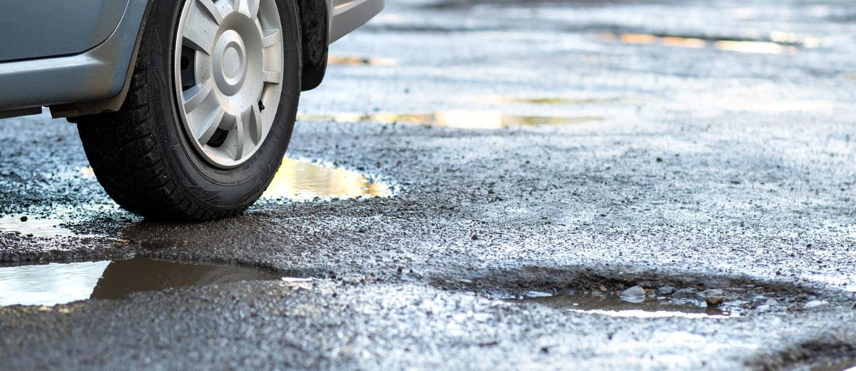 A close-up shot of a car tire driving over potholes filled with water and loose gravel.