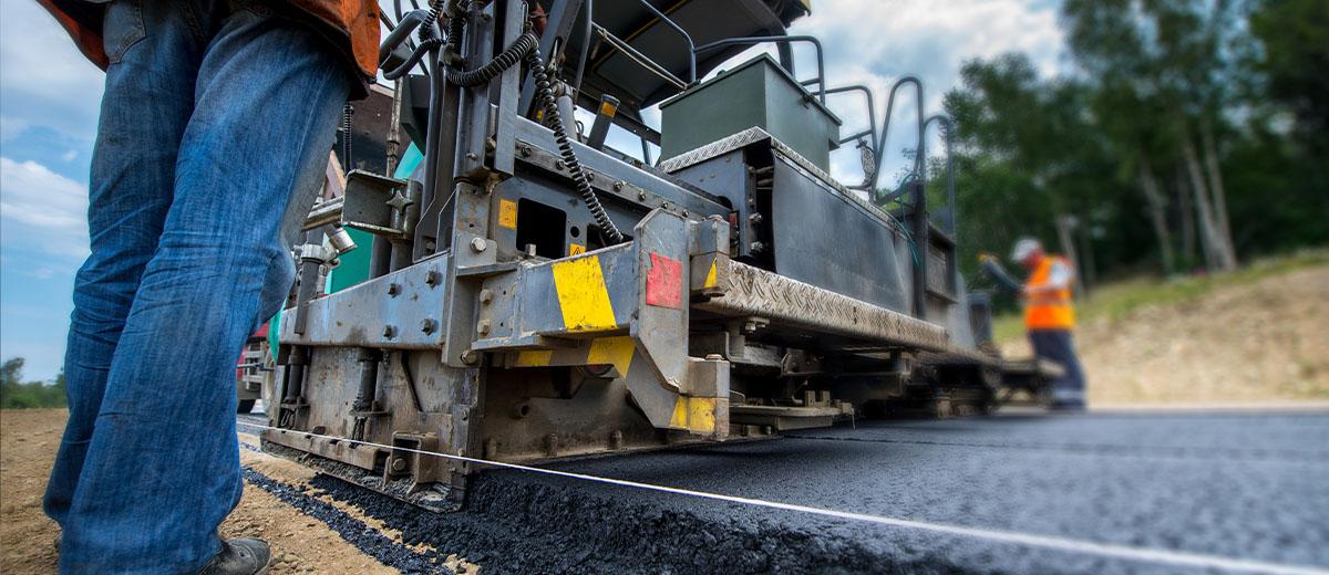 An asphalt paver is being used to smooth out a road, and is being operated by a road worker.